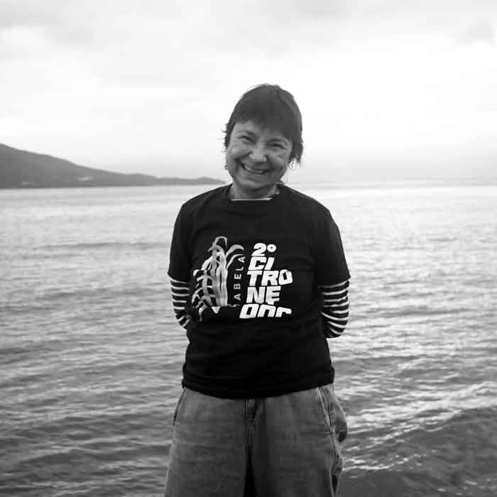 Sou uma mulher cis, branca, de 70 anos, cabelos lisos e curtos. Estou de costas ao canal de São Sebastião com o perfil da Serra do mar ao fundo, visto uma camiseta do Festival e estou sorrindo.