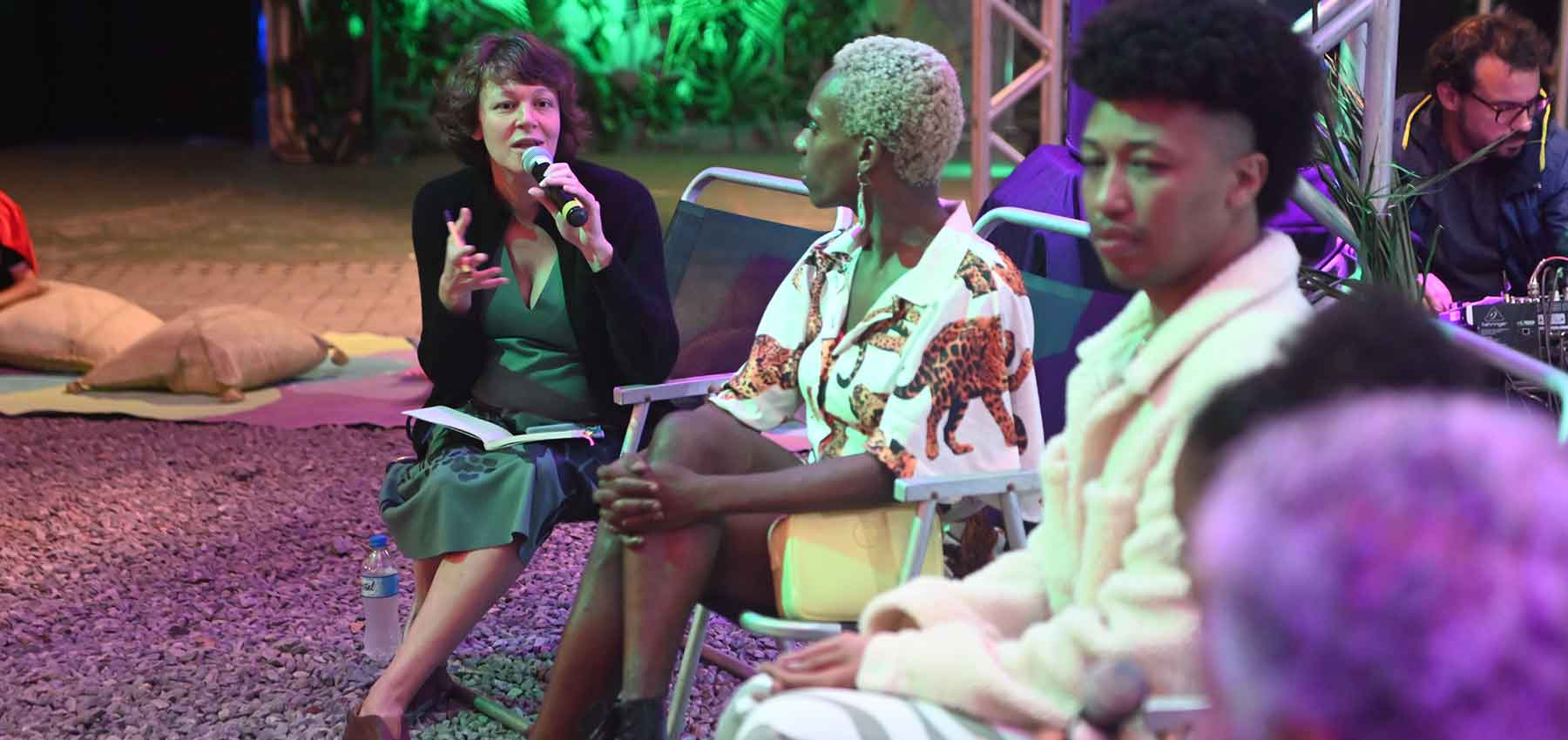 Foto 6 de 6. A imagem é uma foto que mostra um grupo de pessoas sentadas em um ambiente que parece ser um evento, um debate. À direita, uma mulher branca de cabelo escuro, usando vestido verde e um casaco preto, ela segura um microfone, aparentemente falando. Ao lado dela, há outras pessoas sentadas, incluindo uma pessoa negra com cabelo loiro platinado curto e outra com cabelo escuro estilo afro. O ambiente tem uma iluminação colorida, com luzes verdes ao fundo e ao ar livre, com um chão coberto de pedrinhas tipo brita