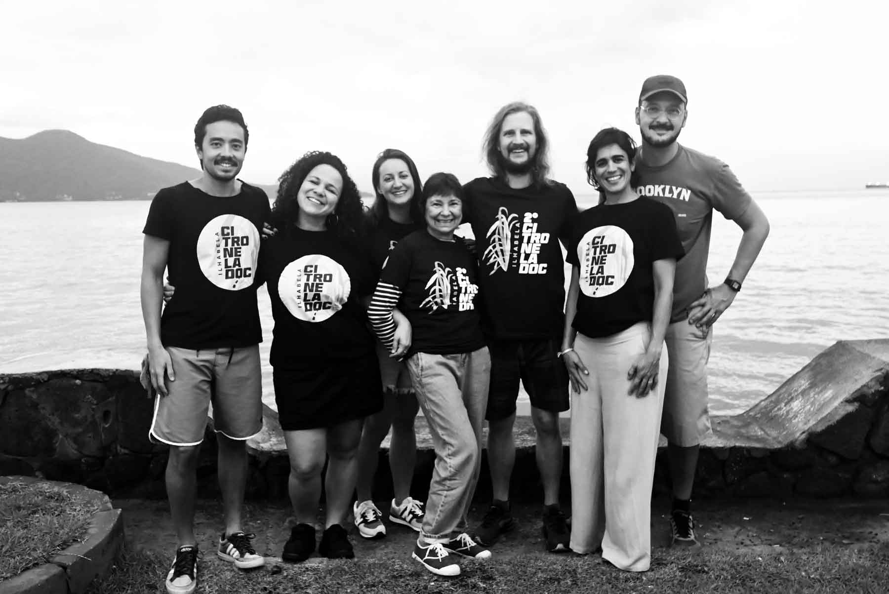 Os sete integrantes do Coletivo Citronela estão posando para a foto, sorrindo, vestindo camisetas do Citronela Doc em frente ao mar. São três homens e quatro mulheres.